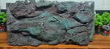 A large rectangular artificial rock backdrop panel with a highly textured, rugged surface, shown wrapped in protective plastic on a wooden shelf.